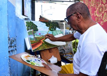 Na fotografia temos um homem negro, usando óculos, pintando uma paisagem em uma tela